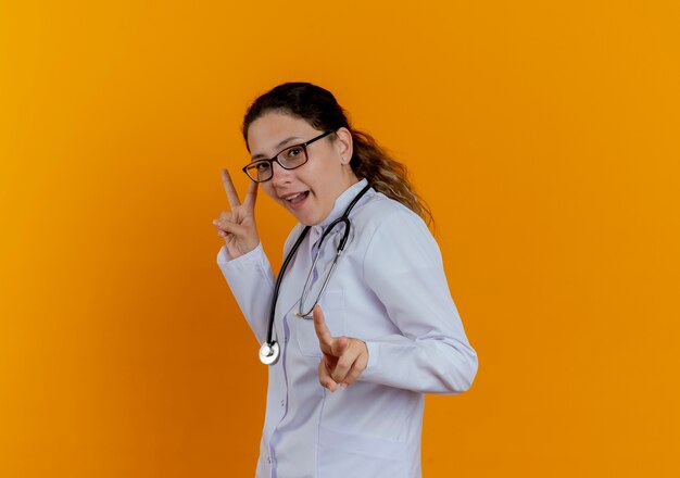 Joyful young female doctor wearing medical robe and stethoscope with glasses showing peace gestures isolated