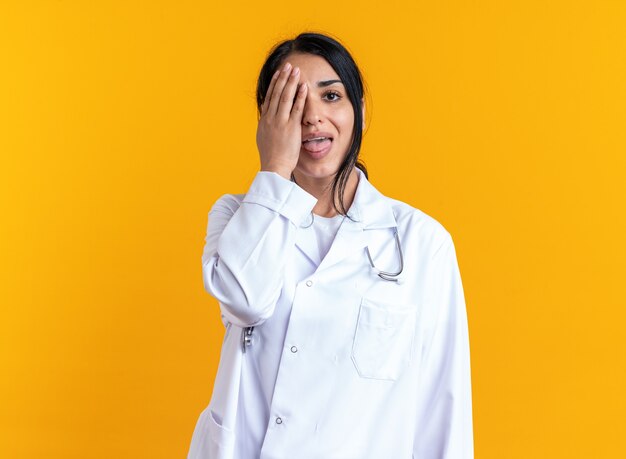 Joyful young female doctor wearing medical robe with stethoscope covered eye with hand isolated on yellow wall