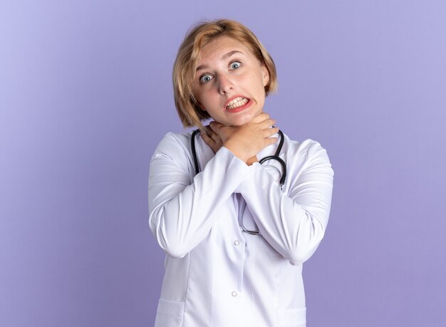 Joyful young female doctor wearing medical robe with stethoscope showing suicide gesture isolated on blue wall