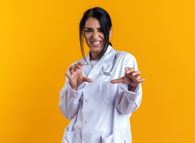 Joyful young female doctor wearing medical robe with stethoscope showing tiger style gesture isolated on yellow wall