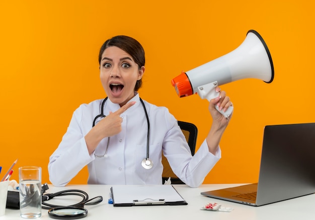 Free photo joyful young female doctor wearing medical robe with stethoscope sitting at desk work on computer with medical tools holding and points to loudspeaker on isolation yellow wall