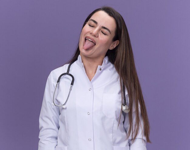 Joyful young female doctor wearing medical robe with stethoscope stucks out tongue