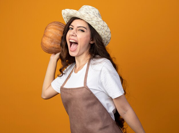 Joyful young female gardener in uniform wearing gardening hat holds pumpkin isolated on orange wall with copy space