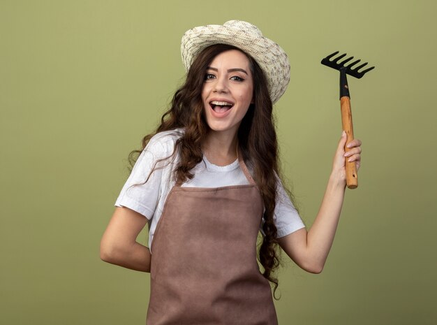 Joyful young female gardener in uniform wearing gardening hat holds rake isolated on olive green wall