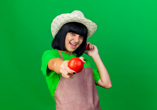 Il giovane giardiniere femminile allegro in uniforme che porta il cappello di giardinaggio tiene i pomodori