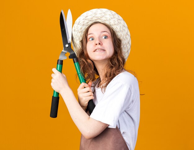 Gioiosa giovane giardiniere femmina che indossa cappello da giardinaggio tenendo la forbice da potatura isolata sulla parete arancione