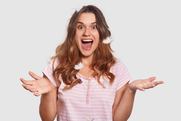 Joyful young female has onjoyed expression, plays with feathers in bedroom, dressed in comfortable pyjamas, has playful look, isolated on white. Rest and lifestyle concept