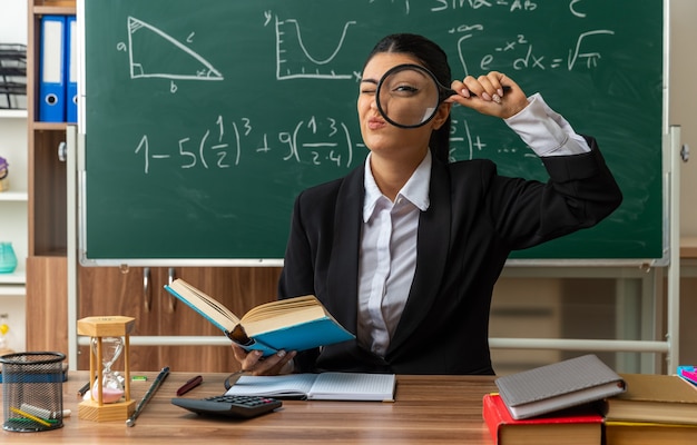 Gioiosa giovane insegnante si siede al tavolo con materiale scolastico guardando davanti con lente d'ingrandimento in classe