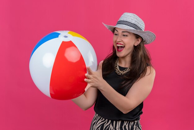 Joyful young female traveler wearing black undershirt in hat holding inflatable ball on red wall