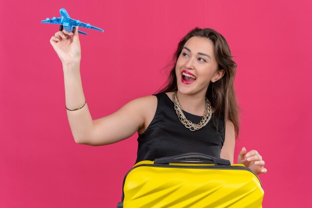 Радостная молодая женщина-путешественница в черной майке играет с игрушечным самолетиком на красной стене