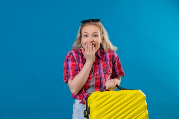 Joyful young female traveler wearing red shirt and glasses on head holding suitcase covered mouth on isolated blue wall