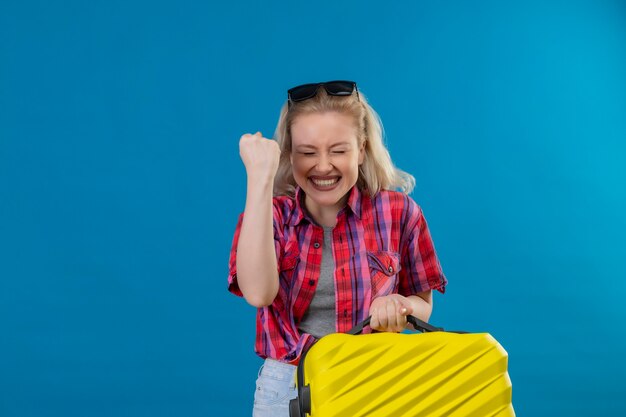 Радостная молодая женщина-путешественница в красной рубашке и очках на голове держит чемодан на изолированной синей стене