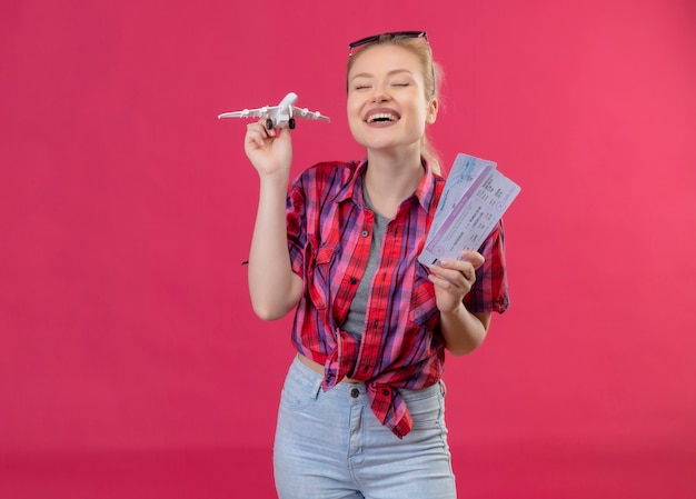 Gioiosa giovane viaggiatrice che indossa la camicia rossa e gli occhiali sulla sua testa tenendo il piano giocattolo con i biglietti sulla parete rosa isolata