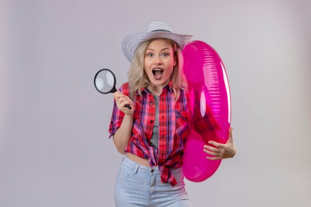 Joyful young female traveler wearing red shirt in hat holding inflatable ring and magnifier on isolated white wall