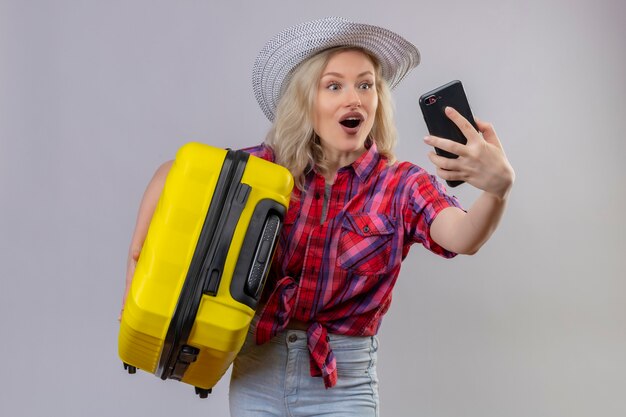 孤立した白い壁に自分撮りを取っているスーツケースを保持している帽子に赤いシャツを着てうれしそうな若い女性旅行者