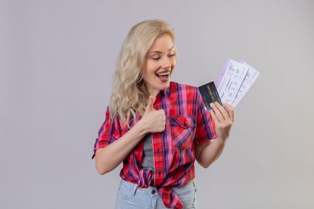 Joyful young female traveler wearing red shirt holding credit card and tickets her thumb up on isolated white wall