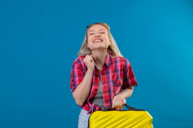 Joyful young female traveler wearing red shirt holding suitcase on isolated blue wall