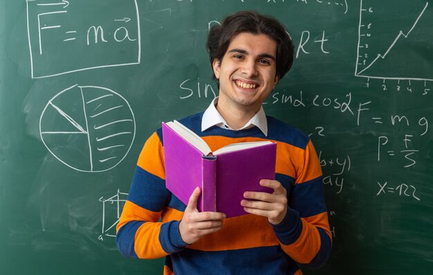 joyful young geometry teacher standing in front of chalkboard in classroom holding book looking at front