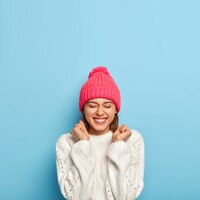 Joyful young girl feels overjoyed, raises clenched fists, being in good mood, wears white sweater and pink hat, dressed in warm clothes during cold autumn day, isolated on blue wall