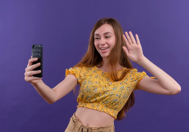 Joyful young girl holding mobile phone and doing hi gesture at mobile phone on isolated purple space