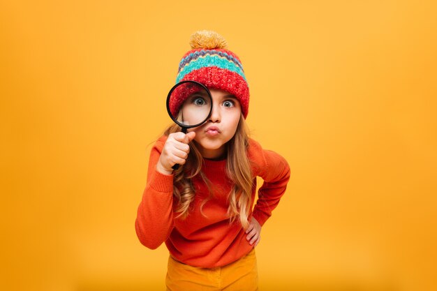 스웨터와 모자에 즐거운 어린 소녀 오렌지 위에 돋보기로 카메라를보고