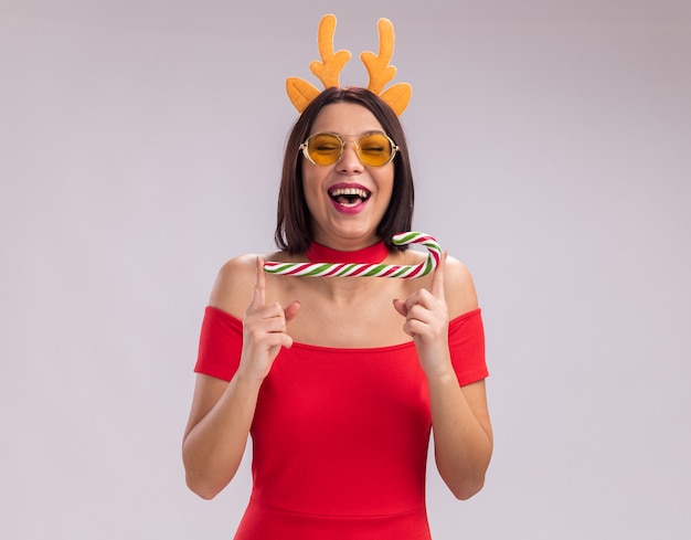 無料写真 トナカイの角のヘッドバンドと白い背景で隔離の目を閉じて笑ってクリスマスクリスマスキャンディケインを保持しているメガネを身に着けているうれしそうな少女