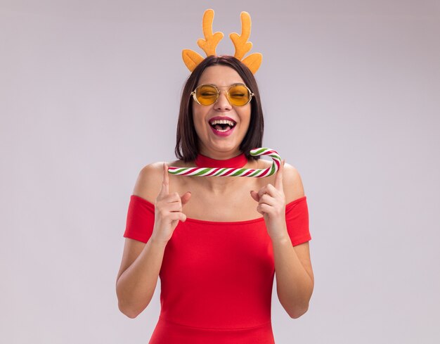 Joyful young girl wearing reindeer antlers headband and glasses holding christmas christmas candy cane laughing with closed eyes isolated on white background