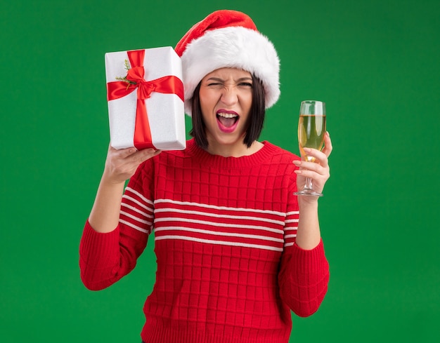 Joyful young girl wearing santa hat holding gift package near head and glass of champagne looking at camera winking isolated on green background