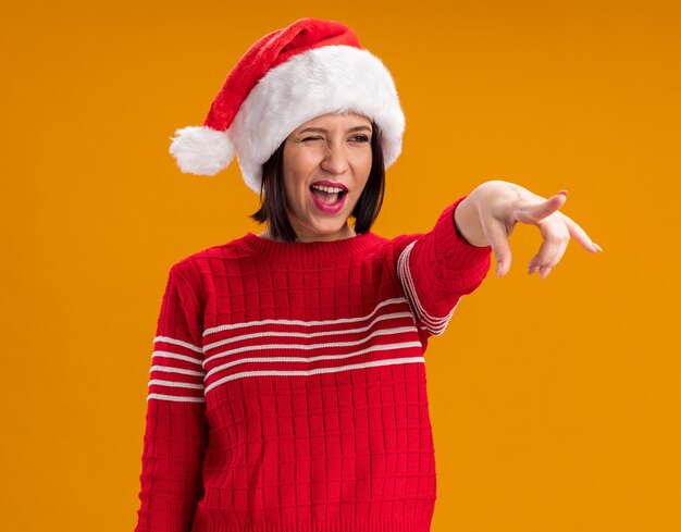 Joyful young girl wearing santa hat looking and pointing at side winking isolated on orange wall