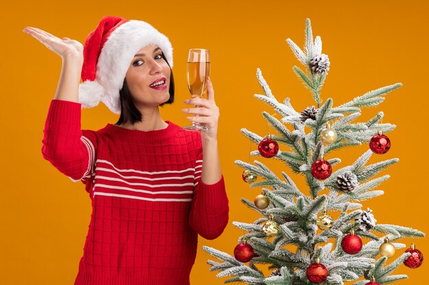 Joyful young girl wearing santa hat standing near decorated christmas tree holding glass of champagne looking at camera showing empty hand isolated on orange background