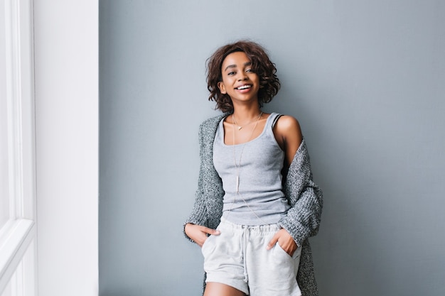 Free photo joyful young girl with short curly hair standing next to gray wall and big white window. wearing casual home clothes, gray cardigan, shirt, shorts, long stylish necklace.