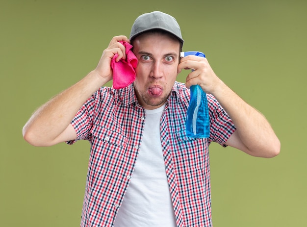 joyful young guy cleaner wearing cap holding cleaning agent with rag around face showing tongue isolated on olive green wall