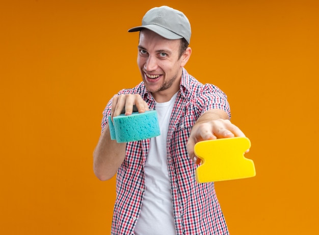 Joyful young guy cleaner wearing cap holding out cleaning sponges at camera isolated on orange background