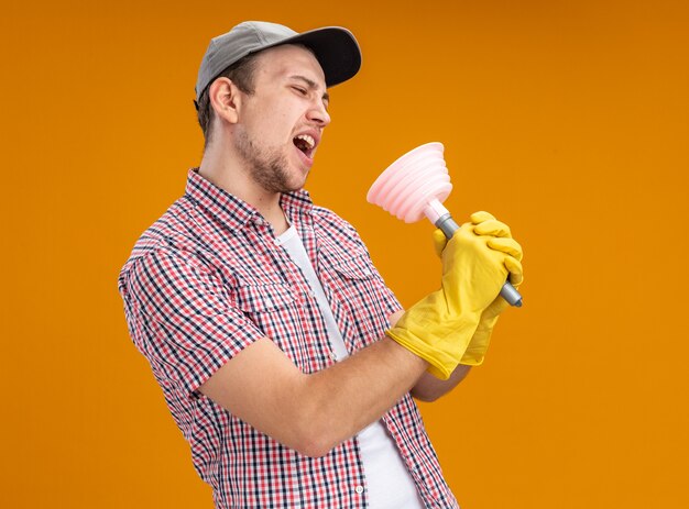 joyful young guy cleaner wearing cap with gloves holding plunger singing isolated on orange wall