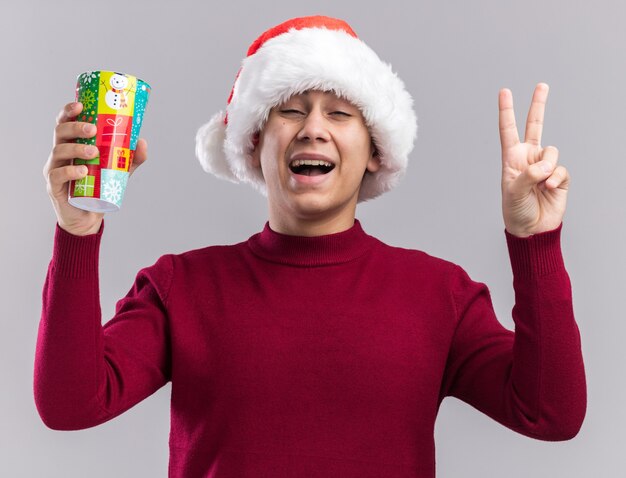 Joyful young guy wearing christmas hat holding christmas cup showing peace gesture isolated on white wall