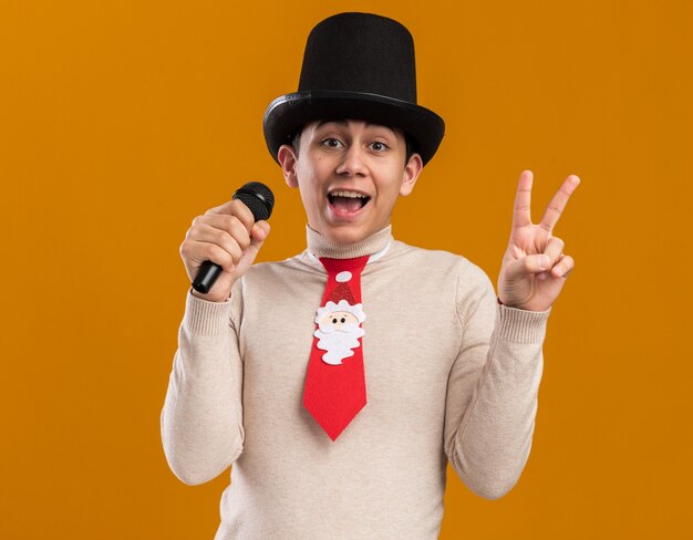 Радостный молодой парень в шляпе с рождественским галстуком говорит в микрофон, показывая жест мира, изолированный на желтой стене