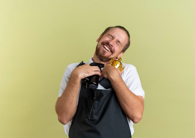 Joyful young handsome barber holding hair clippers, spray bottle and winner cup with closed eyes isolated on olive green