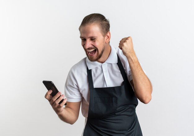 Gioioso giovane barbiere bello che indossa la tenuta uniforme e guardando il telefono cellulare e alzando il pugno isolato su bianco