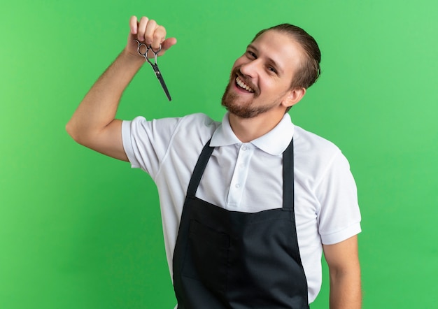 Joyful young handsome barber wearing uniform holding scissors and comb isolated on green