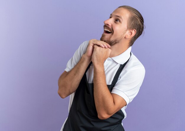 Joyful young handsome barber wearing uniform keeping hands together looking at side isolated on purple