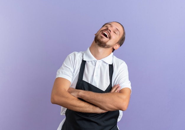 Joyful young handsome barber wearing uniform standing with closed posture and closed eyes isolated on purple