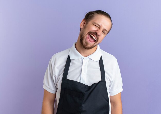 Joyful young handsome barber wearing uniform winking isolated on purple copy space