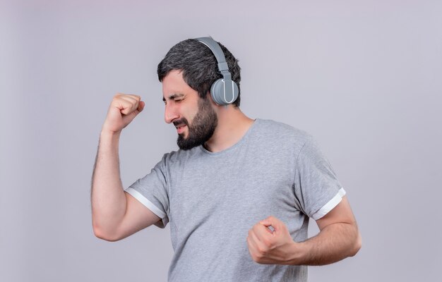 Joyful young handsome caucasian man wearing headphones listening to music with clenched fists and closed eyes isolated on white