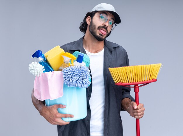 Joyful young handsome cleaning guy wearing t-shirt and cap holding bucket of cleaning tools showing tongue with mop isolated on white wall