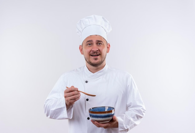 Joyful young handsome cook in chef uniform holding bowl and spoon on isolated white wall with copy space