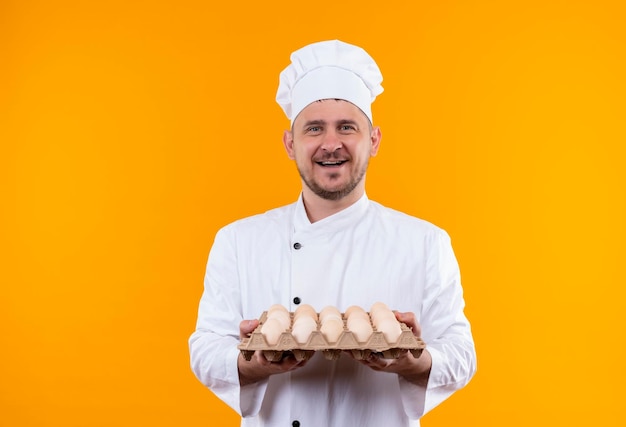 Gioioso giovane e bello cuoco in uniforme da chef che tiene in mano un cartone di uova isolato su una parete arancione