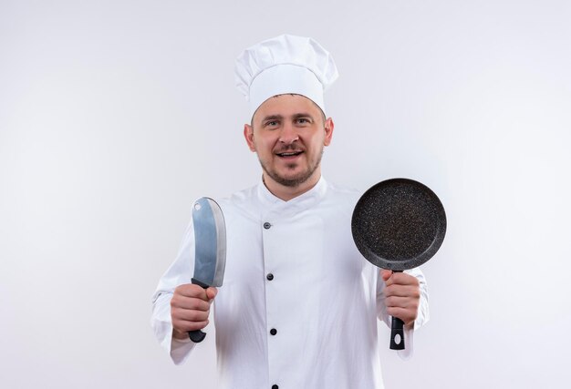 Joyful young handsome cook in chef uniform holding cleaver and frying pan on isolated white wall with copy space