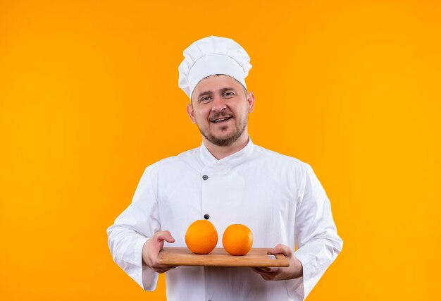 Joyful young handsome cook in chef uniform holding cutting board with oranges on it isolated on orange wall