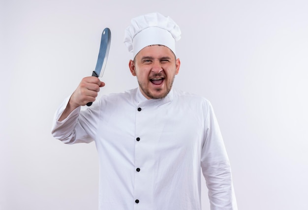 孤立した白い壁にナイフを保持しているシェフの制服を着たうれしそうな若いハンサムな料理人