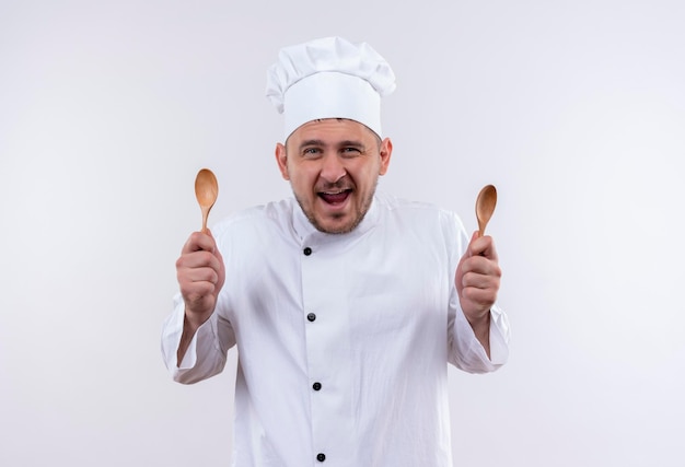 Joyful young handsome cook in chef uniform holding spoons on isolated white wall